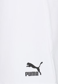 Maglietta sportiva bianca con una texture liscia, caratterizzata da un logo Puma nero nella parte inferiore. Design semplice senza motivi o accenti aggiuntivi.