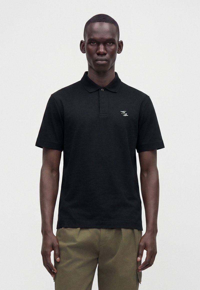 Homme portant un polo noir à manches courtes avec un petit logo blanc sur la poitrine gauche et un pantalon vert olive, debout devant un fond uni.