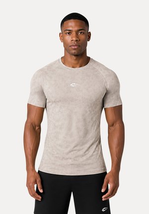 Un uomo è in piedi, rivolto in avanti, indossa una camicia aderente color beige chiaro a maniche corte e pantaloncini neri con un piccolo logo bianco su ciascuno.