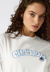 T-shirt en coton blanc avec un logo "CHAMPION" bleu de style bulle et un petit graphisme de personnage de dessin animé. Texture douce, coupe normale.