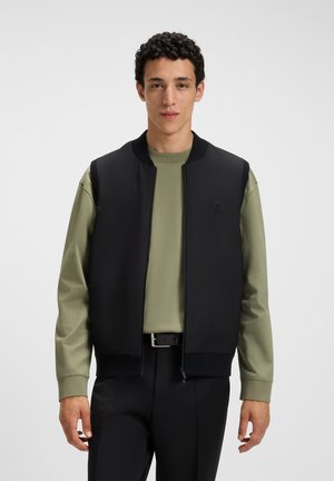 Giovane uomo che indossa un gilet nero sopra una camicia a maniche lunghe verde oliva e pantaloni neri, in piedi di fronte a uno sfondo chiaro e uniforme.