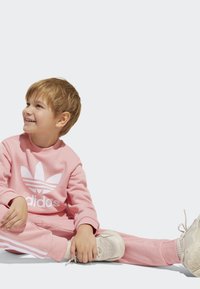 Svetloružový Adidas sveter a tepláky s bielym logom a pruhmi. Svetlé tenisky dopĺňajú outfit. Dieťa sedí s rukami na topánkach.