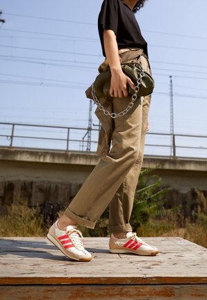 Person trägt beige Hose und weiße Sneakers mit roten Streifen, hält eine olivgrüne Tasche mit einer großen silbernen Kette, steht im Freien.