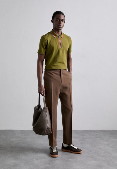 Camisa polo verde con cremallera y cuello multicolor, combinada con pantalones marrones ajustados y zapatillas negras. Lleva un bolso de cuero marrón.