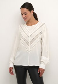 Blouse blanche à manches longues présentant un motif en chevron avec un délicat détail en dentelle et des poignets froncés. Tissu lisse et léger.