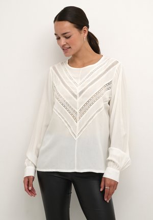 White long-sleeve blouse featuring a chevron design with delicate lace detailing and gathered cuffs. Smooth, lightweight fabric.