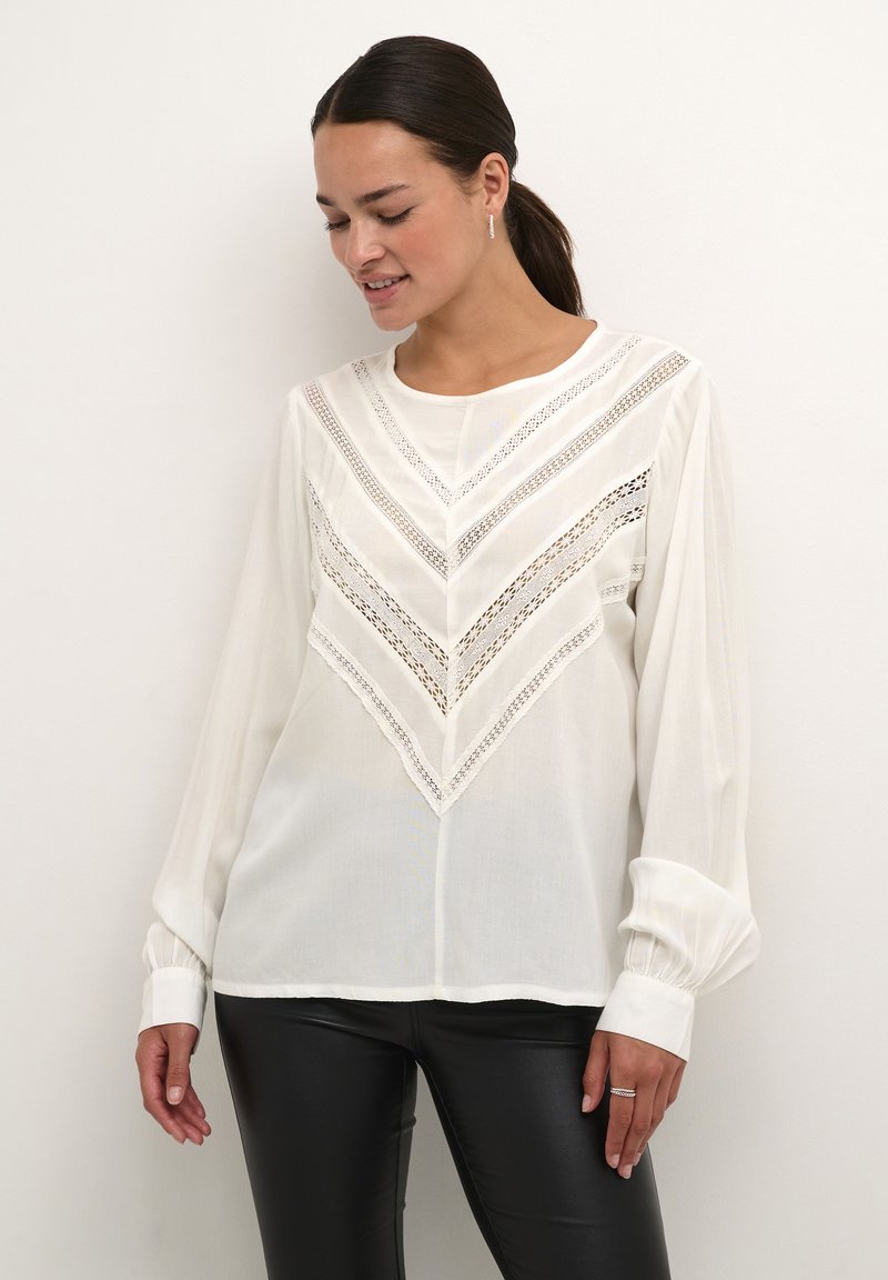 Blouse blanche à manches longues présentant un motif en chevron avec un délicat détail en dentelle et des poignets froncés. Tissu lisse et léger.