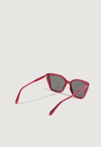 Gafas de sol cat-eye rojas translúcidas con lentes oscuros, abiertas y descansando sobre una superficie gris clara, vistas desde la parte trasera.