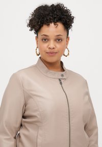 Μπεζ ψεύτικο δερμάτινο μπουφάν με όρθιο γιακά, μπροστινό φερμουάρ και διακριτές ραφές. Το μοντέλο φοράει κρίκους στα αυτιά.