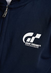 Felpa con zip di colore blu navy realizzata in tessuto morbido, caratterizzata da un grande logo bianco di Gran Turismo sulla parte sinistra del petto.