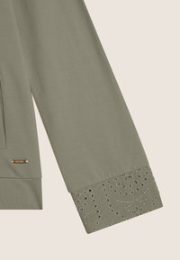 Sudadera color caqui con manga de construcción sin costuras, bordado texturizado en el puño y un pequeño logo de metal "Freddy" como acento.