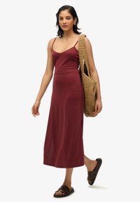 Vestido midi burdeos ajustado con tirantes finos, de textura suave y con una abertura lateral. Combinado con un bolso tejido en tono marrón claro y sandalias deslizantes marrones.