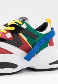 Zapatilla colorida que presenta una mezcla de materiales en rojo, verde, negro y blanco. Destacan los cordones amarillos y una cinta azul que acentúa el talón.