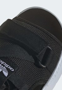 Close-up of a black Adidas sandal with mesh fabric, adjustable strap, white logo, and white sole detailing.