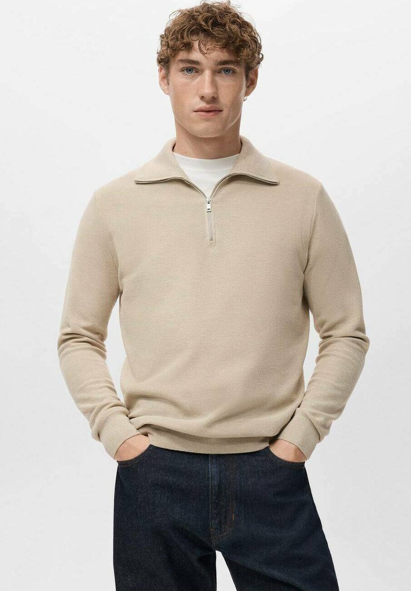 Beige pullovertrui met een halve ritskraag, gestructureerde stof, lange mouwen en geribbelde boorden, gedragen over een wit shirt met ronde hals.