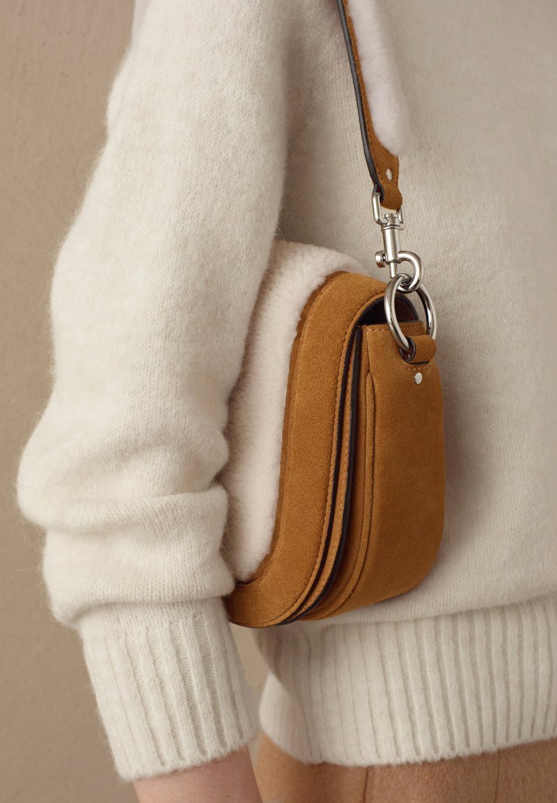 Manica di un maglione bianco in lana con polsino a coste accanto a una piccola borsa a spalla in suede beige con hardware in metallo argentato, indossata su uno sfondo beige.