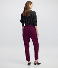 Pantalones tapered en color marrón oscuro con cintura alta, bolsillos traseros y una textura suave, combinados con una camisa negra de manga larga y tacones.