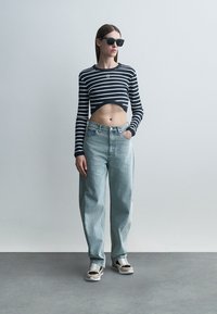 Kvinne iført svarte solbriller, svart-hvitt stripet kortet langermet topp, lyseblå høytlivs jeans og joggesko, står med hånden i lommen.