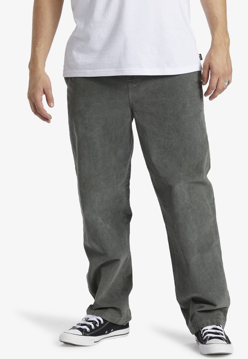 Pantalones de pana gris con un ajuste relajado y costillas verticales sutiles; combinados con zapatillas de lona negras con suelas y cordones blancos.