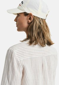 Witte baseballpet met rode tekst, gedragen met een licht gestreept overhemd met knopen, met de nadruk op de textuur van de stof en de strakke lijnen.