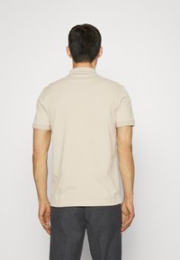 Homme de dos portant un polo à manches courtes beige et un pantalon gris foncé devant un fond blanc uni.