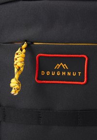 Czarny plecak z tkaniny z czerwoną i złotą naszywką logo "DOUGHNUT". Posiada żółty pleciony uchwyt suwaka i fakturowaną powierzchnię.