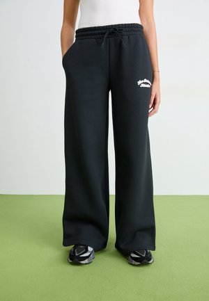 Pantaloni della tuta neri con vita regolabile, design a gamba larga, tasche laterali e testo ricamato bianco sull'anca sinistra. Tessuto liscio e morbido.