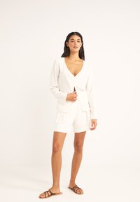 Top de lino blanco con escote en V profundo y mangas largas, combinado con pantalones cortos a juego. Presenta botones visibles y un corte holgado. Sandalias marrones.