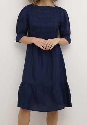Femme portant une robe bleu marine avec un corsage smocké, des manches ballon jusqu'au coude et un ourlet à volants, debout devant un fond blanc uni.