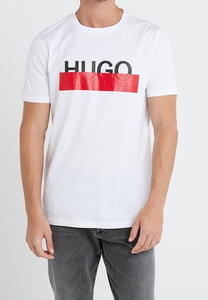 Mężczyzna w białym t-shircie z krótkim rękawem z czarno-czerwonym logo Hugo Boss na piersi i ciemnoszarych dżinsach.