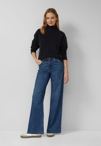 Schwarzer Rollkragenpullover mit geripptem Kragen, kombiniert mit weit geschnittenen blauen Jeans. Das Outfit wird durch einen schwarzen Gürtel und beige Sneaker ergänzt.