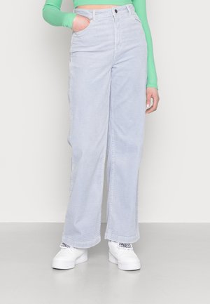 Pantalones de pana de tiro alto y pierna ancha en azul claro con un acabado texturizado, cinco bolsillos y cierre frontal con botón. Zapatillas blancas abajo.