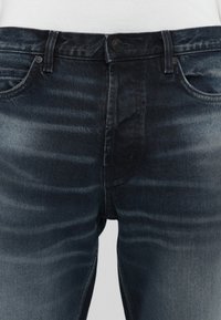 Jeans de mezclilla azul oscuro con cierre de botón, cinco bolsillos y un leve desvanecimiento a lo largo de los muslos, que proporciona un aspecto texturizado.