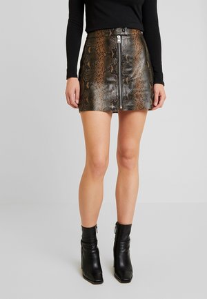 Woman wearing a brown and black snakeskin mini skirt with front zipper, black long-sleeve top, and black ankle boots in a neutral setting.