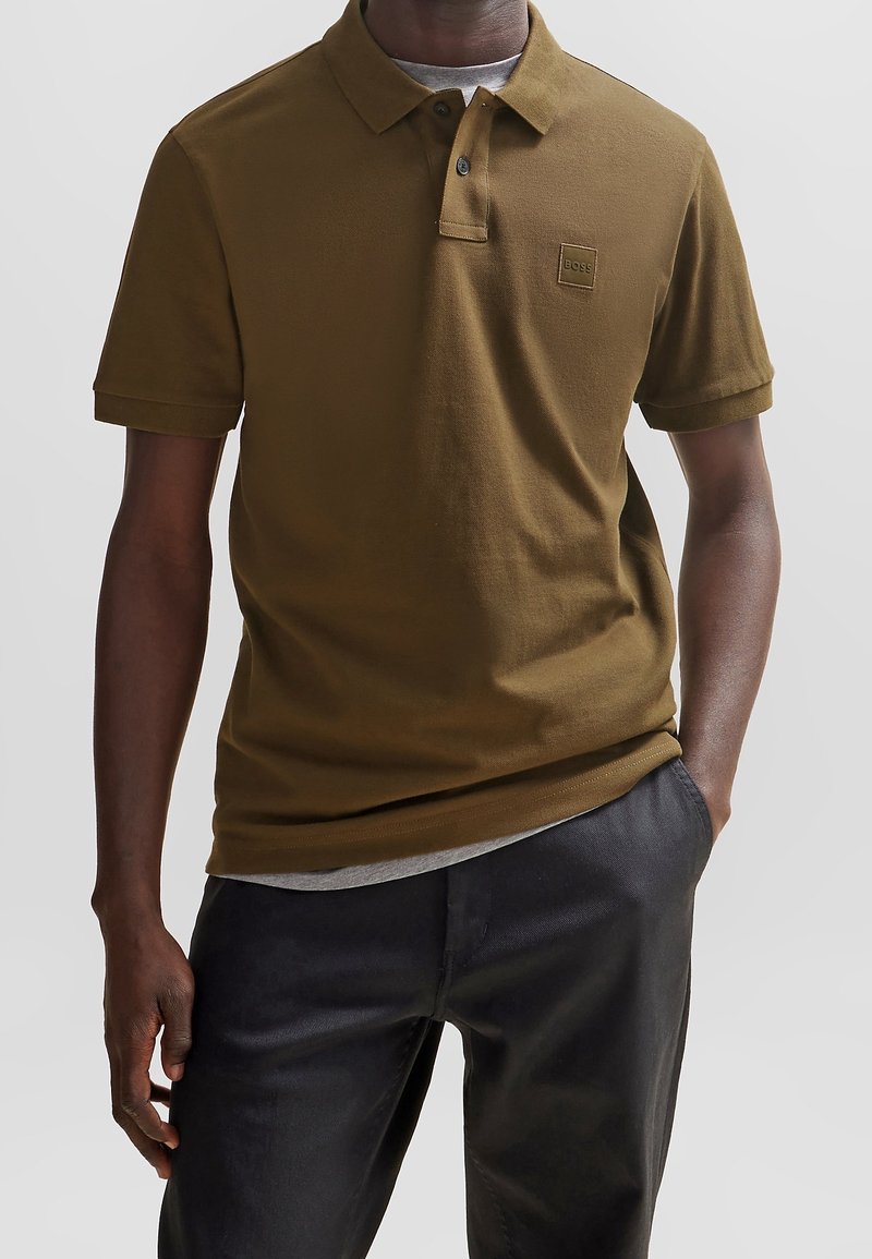 Polo marron en tissu texturé, avec une patte de boutonnage à trois boutons et un petit écusson logo sur la poitrine, associé à un pantalon foncé ajusté.