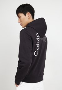 Černá mikina s kapucí a bílým logem "Calvin Klein" na zádech. Vyrobená z bavlny, má uvolněný střih a žebrované manžety.