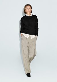 Maglione nero a maglia sopra una camicia bianca con colletto, abbinato a pantaloni beige larghi e scarpe nere. Il modello sta in piedi con le mani in tasca.