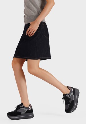 Donna che indossa un top senza maniche grigio, una gonna di denim scuro e sneakers con piattaforma nero-grigio mentre cammina su uno sfondo bianco uniforme.