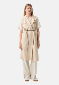 Beige Woll-Wickelweste mit breiten Revers, vorderen Taschen und einem braunen Bindegürtel, kombiniert mit cremefarbener, weit geschnittener Hose und beigen Plateauschuhen.
