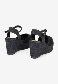 Sandalias cuña negras con una correa para el tobillo de tela texturizada, parte superior lisa y plataforma de yute trenzado. Punta redondeada y cierre de hebilla ajustable.