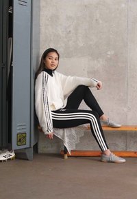 Suéter branco e fofinho com riscas pretas, combinado com leggings pretas que apresentam riscas brancas. Sapatos cinzentos, sentado num banco perto de cacifos.