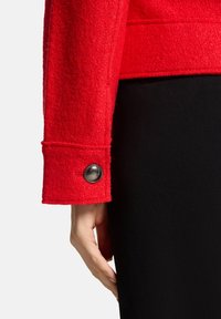 Rode wolmix jas met een mouw die een afgeronde zwarte knoopdetail heeft. De manchetten zijn zorgvuldig afgewerkt met een gladde afwerking. Zwarte stof aan de binnenkant.