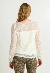 Blusa blanca de encaje con mangas largas y transparentes, que presenta un patrón texturizado y un respaldo sólido. Combinada con leggings marrones de estampado de leopardo.
