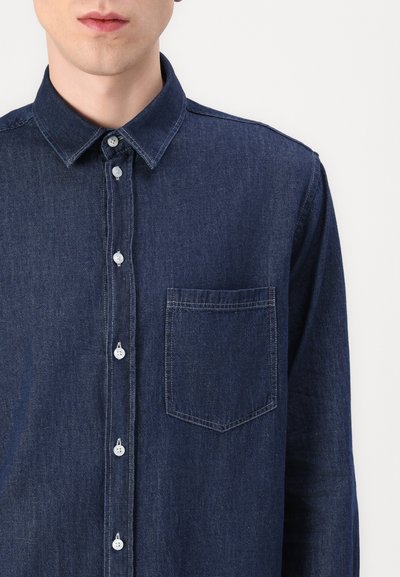 Chemise en denim bleu foncé avec un devant à boutons, col pointu et poche poitrine unique. Présente des boutons blancs et un tissu texturé.