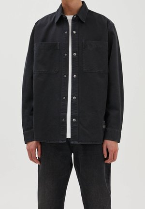 Homme portant une veste-chemise noire à boutons avec deux poches poitrine sur un t-shirt blanc et un pantalon noir sur fond uni.