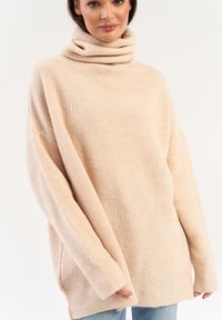 Beżowy, oversize sweter z golfem o prążkowanej fakturze, z długimi r�ękawami i wysokim kołnierzem, w połączeniu z jasnoniebieskimi dżinsami.