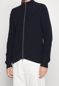 Pletený tmavě modrý svetr na zip s vysokým límcem, žebrovanou texturou, dlouhými rukávy a stříbrným zipem podél přední části.