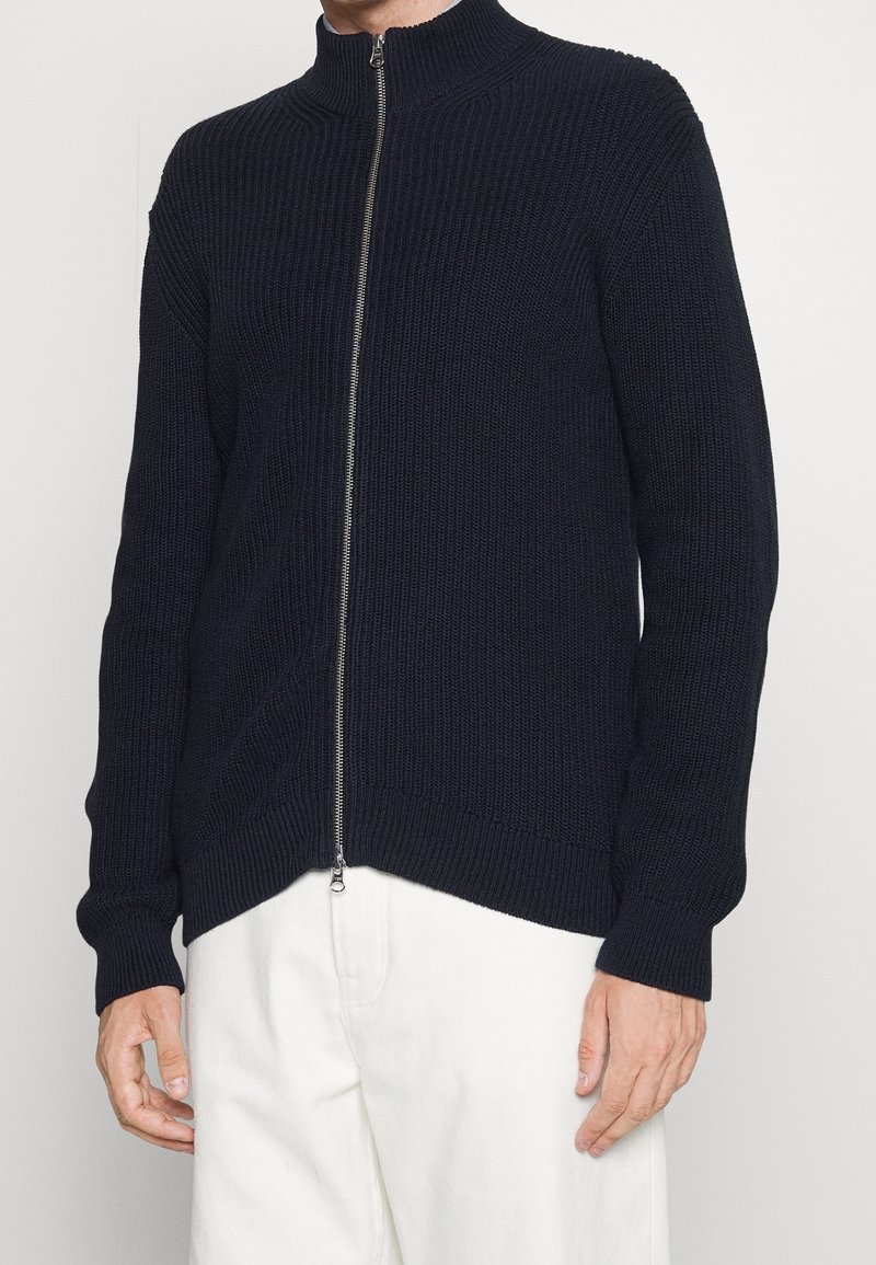 Pletený tmavě modrý svetr na zip s vysokým límcem, žebrovanou texturou, dlouhými rukávy a stříbrným zipem podél přední části.