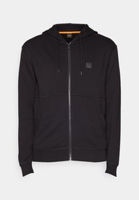 Svart zip-up hoodie med dragsko i huven, featuring ett fyrkantigt patch-logotyp på bröstet och kontrasterande sömmar. Mjuk, bomullsblandning material.