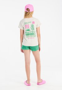 Camiseta color crema con gráficos verdes y rosas en la parte de atrás. Combinada con pantalones cortos verdes y zuecos rosas, con mangas dobladas.